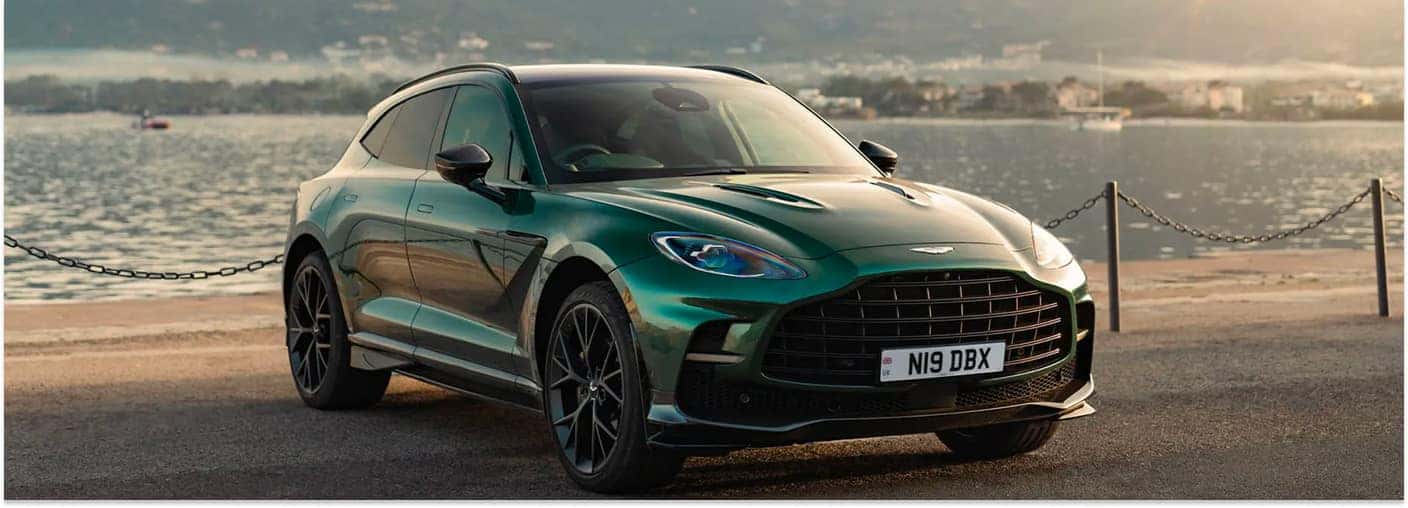 Front three-quarter view of a green 2026 Aston Martin DBX SUV parked along a waterfront promenade.