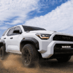 White 2025 Toyota 4Runner kicking up dust on an off-road trail, showcasing its rugged new design, high ground clearance, and bold front grille under a partly cloudy sky.