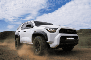 White 2025 Toyota 4Runner kicking up dust on an off-road trail, showcasing its rugged new design, high ground clearance, and bold front grille under a partly cloudy sky.