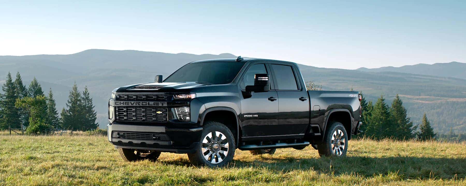 Black Chevy Silverado 2500HD on the grass parked in front of trees and mountains.