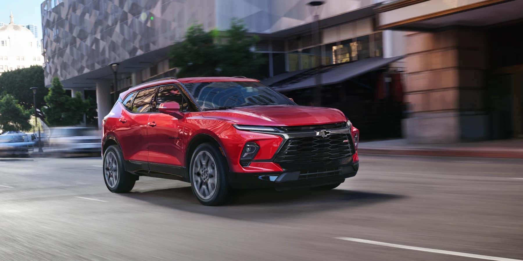 A red 2023 Chevrolet Blazer driving on city streets with buildings and cars in the background. 
