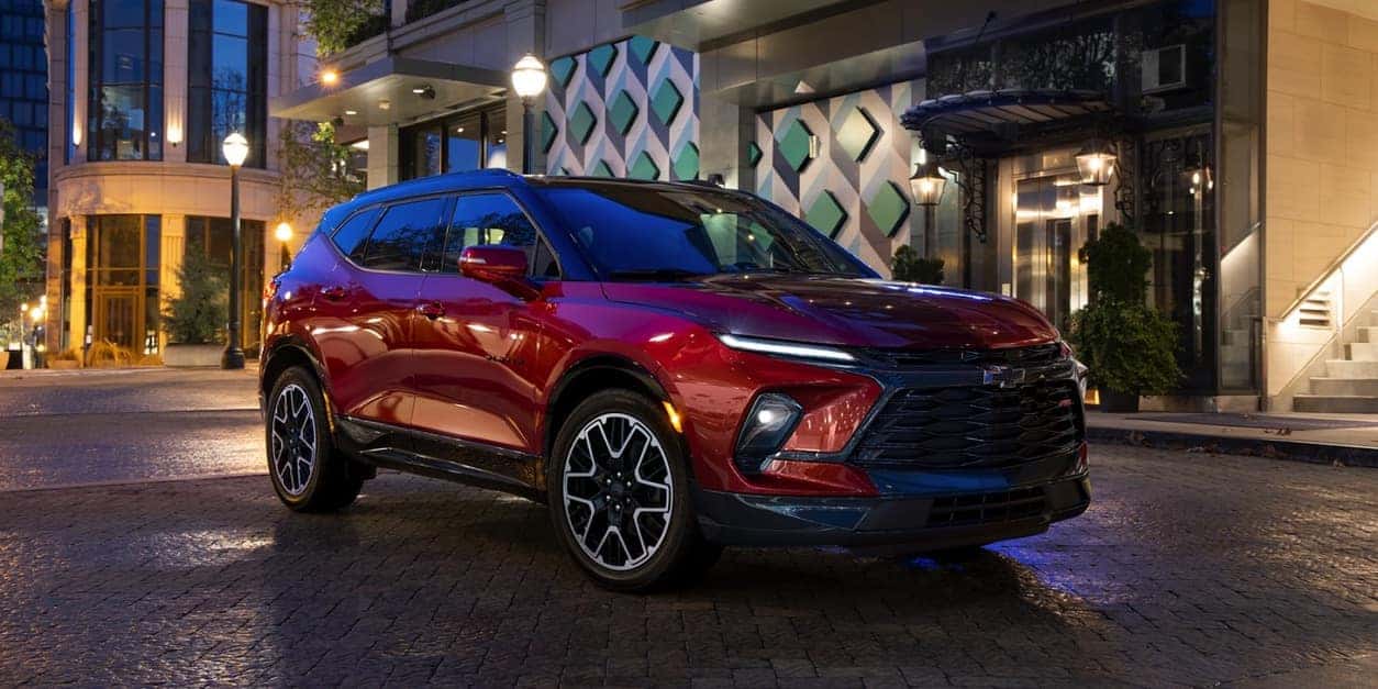 A red 2023 Chevrolet Blazer parked in front of a well-lit building with its headlights turned on.