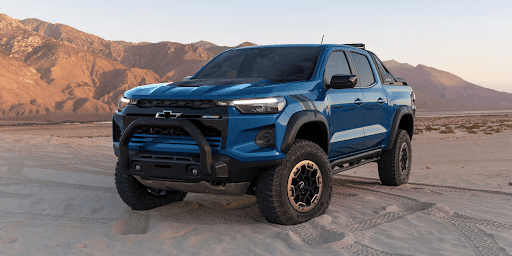 A dark blue 2023 Chevy Colorado on desert land with mountains in the background.