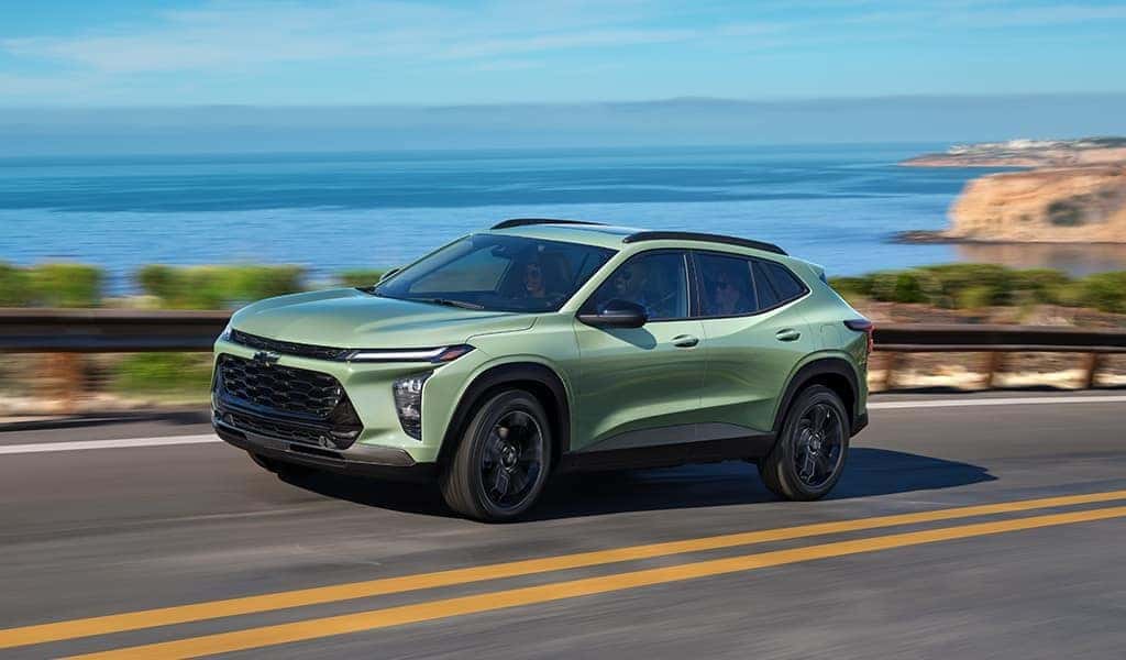 A light green 2024 Chevy Trax driving on the road with a beach in the background.