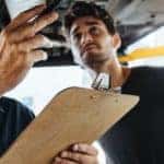 A mechanic with a clipboard is inspecting a car on a lift.