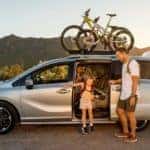A father is opening the rear door for his daughter in a silver 2021 Honda Odyssey Elite with bikes on the roof.