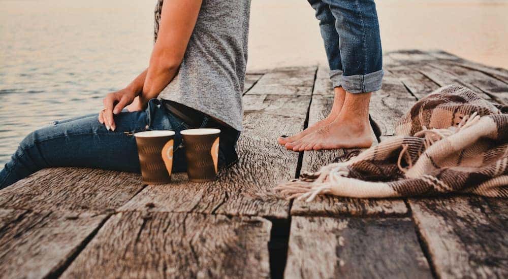 Close up of a man's feet standing behind a woman sitting on a dock with two coffees