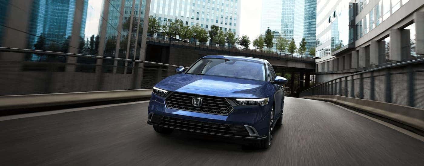 A blue 2023 Honda Accord Sport Hybrid is shown from a front angle driving on a city highway.