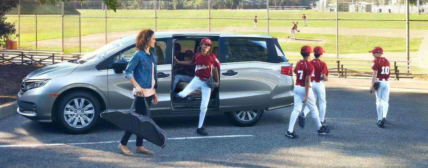 Mom and kids in baseball uniforms climbing out of a silver 2021 Honda Odyssey Elite at a little league field