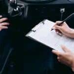 Close-up of a person writing on a clipboard inside a car, showing hands and a gear shift.