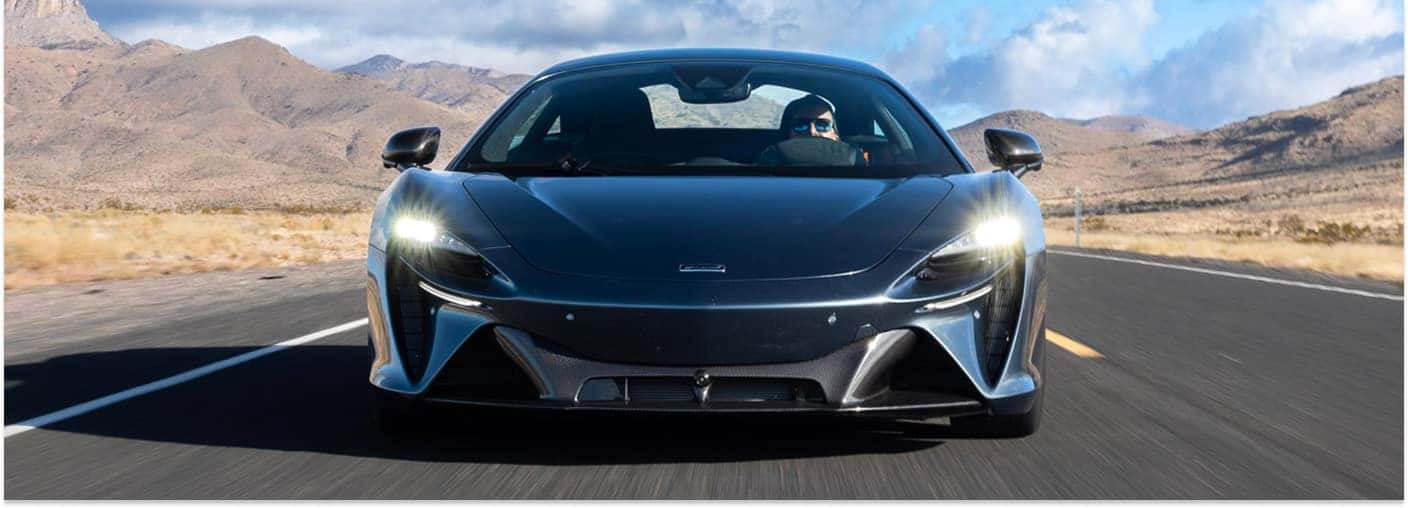 Front view of a dark gray 2026 McLaren Artura driving on an open road with mountains in the background.