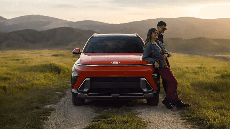 A red Hyundai Kona parked on a scenic dirt road at sunset, with two people leaning against the front enjoying the view.